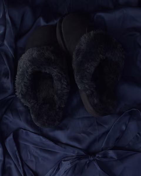 The Elysian in Midnight, charcoal shearling slippers resting on navy satin